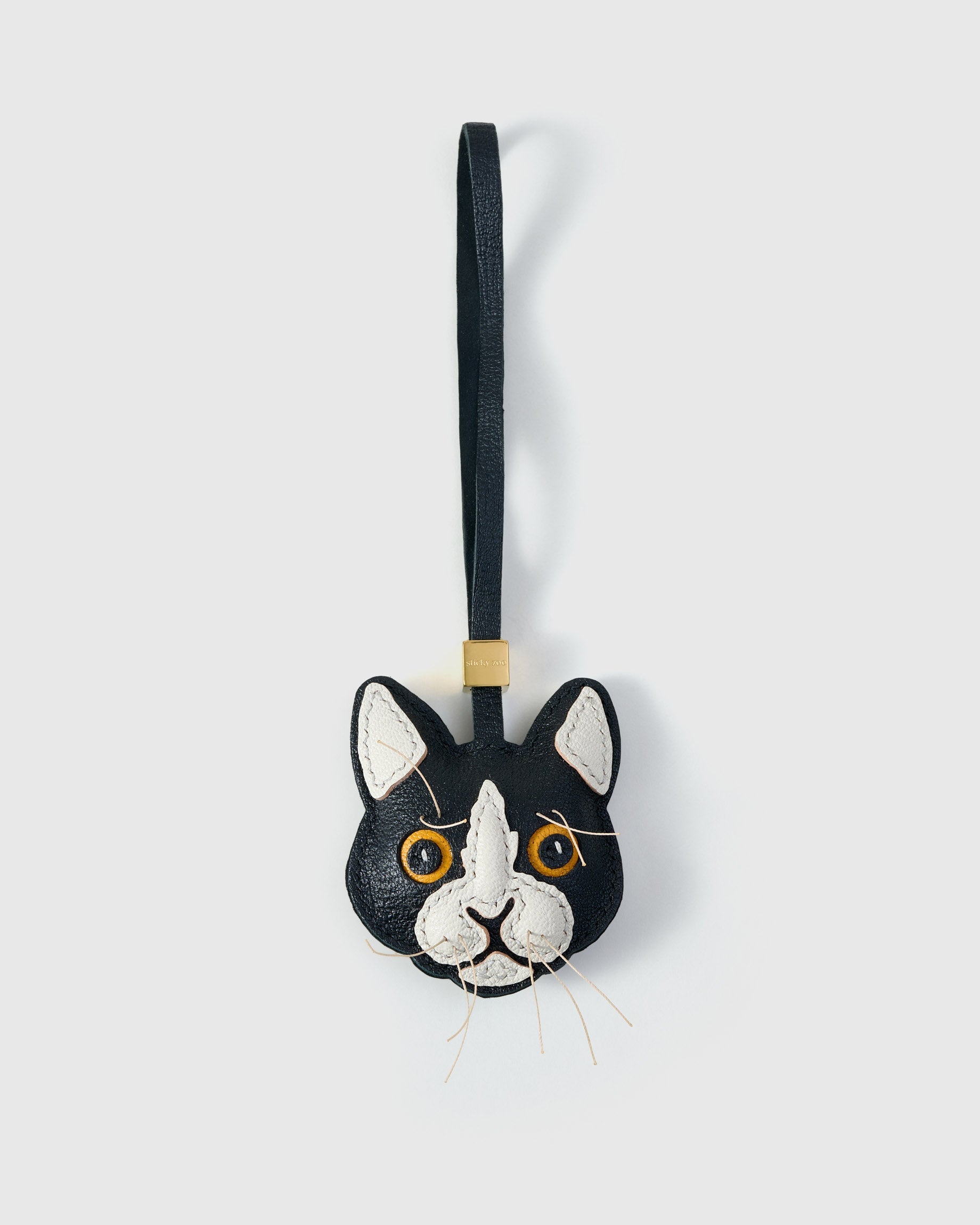 Tuxedo cat charm handmade from French Alran genuine goatskin — black body, white face mask and chest, pink inner ears, gold-yellow eyes, real fiber whiskers, dark navy leather strap. Three days by hand. Arrives in luxury gift packaging. Designed in Amsterdam.