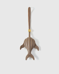 Sculpted taupe whale charm handmade from French Alran genuine goatskin — dimensional 3D body viewed from above, side fins, split tail fluke, cream hand-stitched seams, taupe leather strap. Three days by hand. Arrives in luxury gift packaging. Designed in Amsterdam.