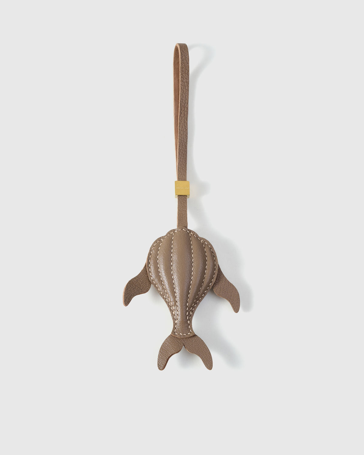 Sculpted taupe whale charm handmade from French Alran genuine goatskin — dimensional 3D body viewed from above, side fins, split tail fluke, cream hand-stitched seams, taupe leather strap. Three days by hand. Arrives in luxury gift packaging. Designed in Amsterdam.