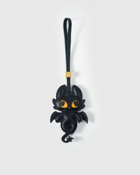 Black dragon charm handmade from French Alran genuine goatskin — deep black body, bat-like wings, large amber-yellow eyes, curled leather tail, black leather strap. Three days by hand. Arrives in luxury gift packaging. Designed in Amsterdam.