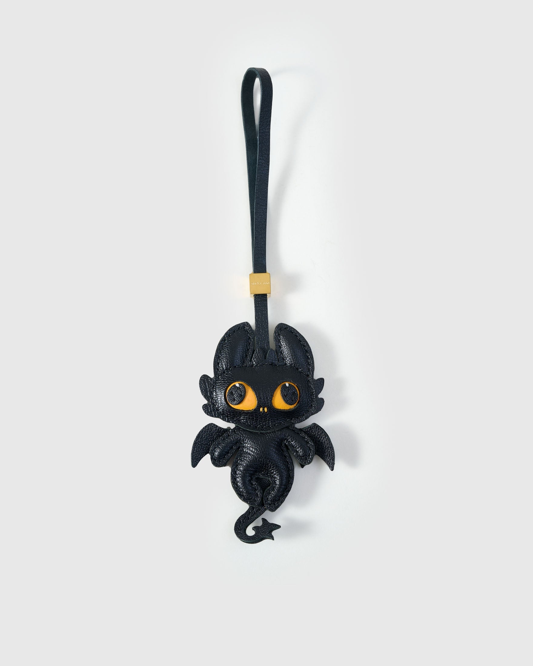 Black dragon charm handmade from French Alran genuine goatskin — deep black body, bat-like wings, large amber-yellow eyes, curled leather tail, black leather strap. Three days by hand. Arrives in luxury gift packaging. Designed in Amsterdam.